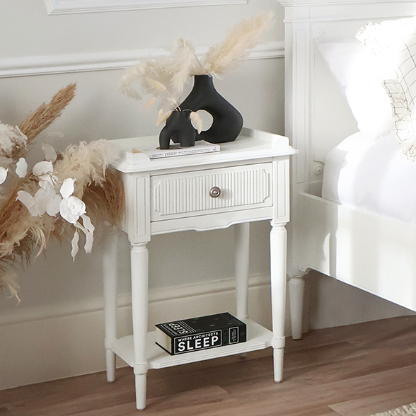 A small, white bedside in a Gustavian style. Styled with a vase on top and to the side of a wooden bed. 