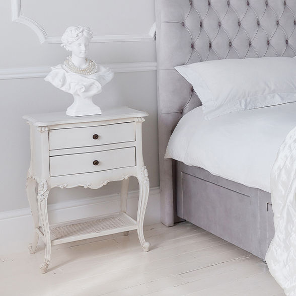 A white bedside table with curved legs and two drawers. Next to an grey upholstered divan bed.